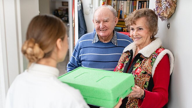 Eine Mitarbeitering des mobilen Menü Service der AWO in Mönchengladbach übergibt einem älteren Paar eine warme Mahlzeit.