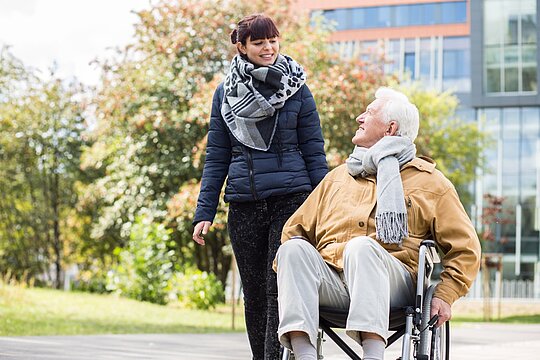 Eine Mitarbeiterin der AWO Mönchengladbach, die hauswirtschaftliche Unterstützung leistet, geht mit einem älteren Mann im Rollstuhl spazieren.